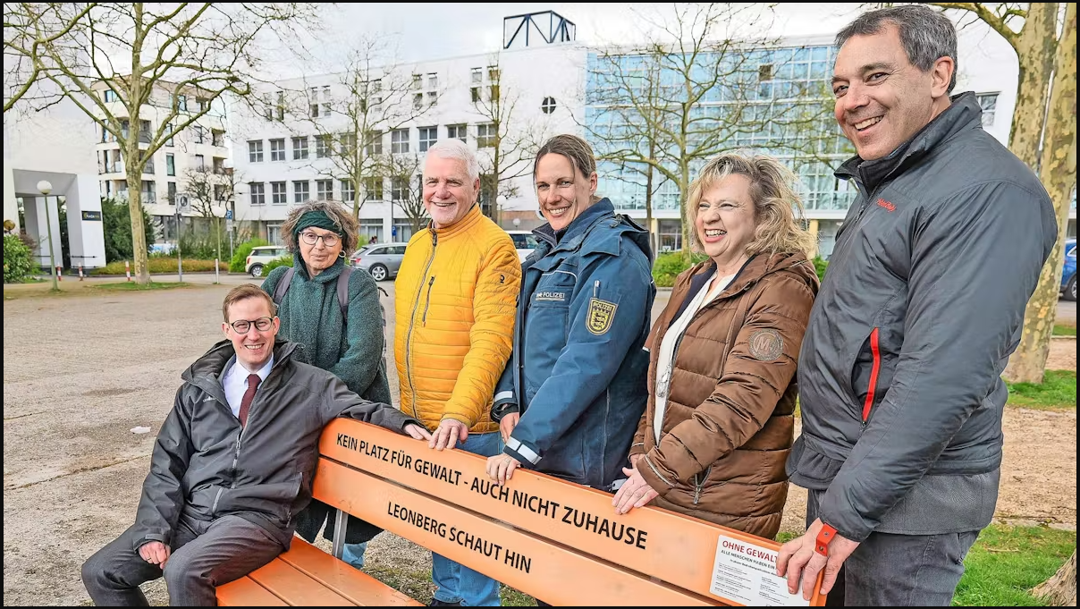 Sechs Personen stehen draußen hinter einer Orangenen Bank mit der Aufschrift "Kein Platz für Gewalt - Auch nicht Zuhause; Leonberg schaut hin"
