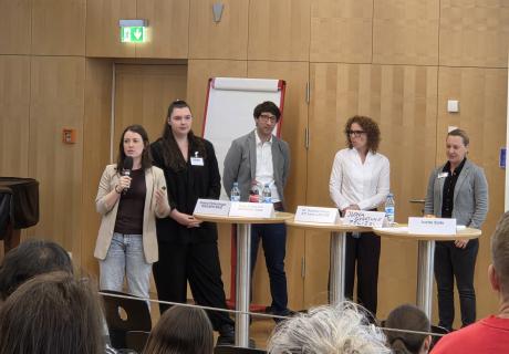 5 Personen stehen vor Stehtischen mit Namensschildern und halten einen Vortrag